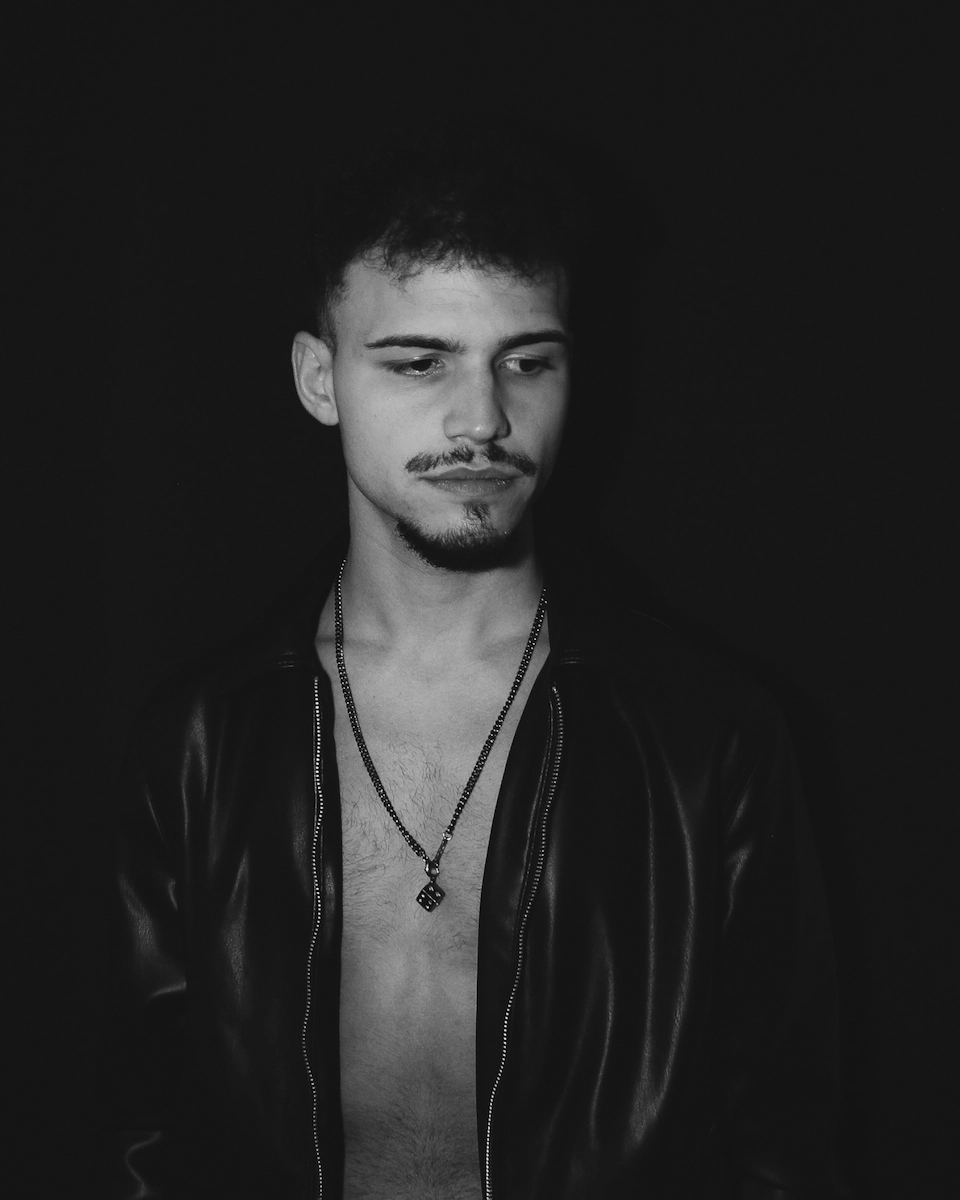 Black-and-white portrait of Kaylor Tyrell, wearing a leather jacket with an open chest and a necklace, gazing downward with a calm expression against a dark background.